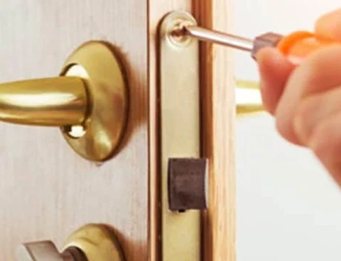 A close-up photograph of a locksmith finishing work on a residential door lock. The light brown wooden door has a newly installed brass-colored lever handle with a round matching backplate on the left side, while a long brass-colored lock plate runs along the door edge. A screwdriver with an orange handle is positioned at the upper screw of the newly installed lock hardware, with the locksmith’s hand holding it as the final screw is being tightened. The rectangular door latch is visible in the center of the lock plate, and part of another brass-colored lock or handle is visible near the lower left corner. The wood door frame is visible along the right edge, with bright light coming through the doorway. The image shows the final stage of installing a replacement residential door lock after the previous lock was no longer functional, representing residential locksmith service in Spring, TX.
