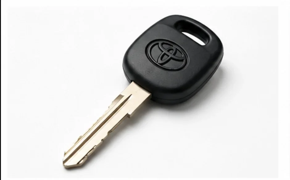 A traditional Toyota car key is displayed diagonally on a clean white background. The key features a black rectangular plastic head with rounded corners and an integrated hole for attaching it to a key ring. The Toyota logo is embossed in the center of the plastic head. Extending from the head is a silver metal key blade with precisely cut grooves and notches designed to match a specific vehicle lock. The key appears clean and unused, with the metal blade reflecting light against the plain white background. The image represents the standard mechanical key commonly used in older Toyota vehicles before remote key fobs and smart key systems became widely available.