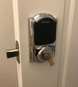 Close-up of a modern electronic smart deadbolt lock with a black touchscreen keypad and chrome finish installed on a white interior door, with a physical key inserted into the lower key cylinder and the door slightly open.