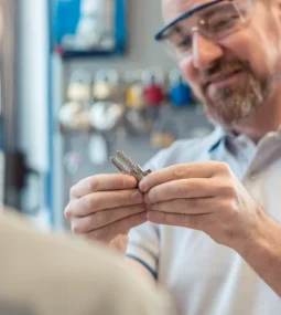Smiling man in protective glasses carefully holding multiple silver keys between his fingers while standing inside a key shop, with another person’s shoulder visible in the foreground and a rack of keys blurred behind him.